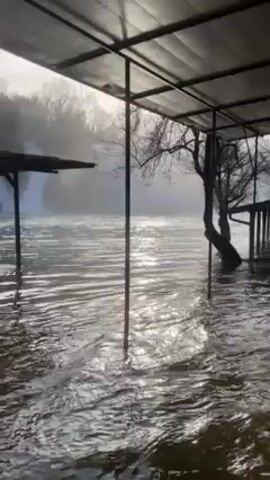 The video shows an area with a waterfall, possibly after a heavy rainfall or flood. The water level is high, and it appears to be flowing under a structure with a roof supported by metal poles. The water is murky, and there is some mist or spray in the air near the waterfall. The video is taken from under the structure, looking out at the scene.