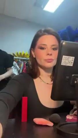 A woman with brown hair and a black top is applying mascara in front of a black makeup mirror. On the table in front of her are a red lipstick tube, a pink makeup sponge, and a round, dark-colored makeup applicator. A child in pajamas walks behind her briefly. The room has clothes and feathered decorations hanging up in the background. A fluorescent light illuminates the scene.