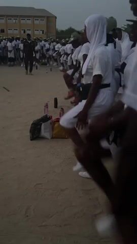 The video shows a group of people dressed in white clothes and green caps, possibly in some sort of training or drill. They are on a dusty, outdoor field, with a building visible in the background. Some individuals are moving and exercising in unison. There are personal belongings like bags and water bottles on the ground near where they are standing.