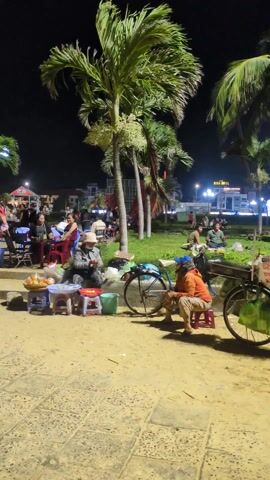 At night, people gather in an outdoor area among palm trees. There are several groups seated at tables, some eating or conversing. A woman walks barefoot across a concrete step, carrying a striped bag. Two people are sitting on small stools near a bicycle, with some food items displayed on a blue stool. The area is illuminated by streetlights and the lights of nearby buildings. The ground is a sandy-colored paving.