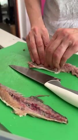A person is shown in a close-up view, with their hands actively engaged in preparing fish on a bright green cutting board. A knife with a metal blade and a white and grey handle is positioned on the cutting board. Two pieces of filleted fish, with bones and flesh visible, are laid out on the board. The person is using their fingers to meticulously work with one of the fish fillets, appearing to remove small bones or excess tissue. The environment suggests a kitchen or food preparation area, with a blurred background that hints at other objects or a wall. The lighting is bright, illuminating the details of the fish and the hands at work.
