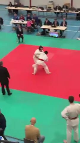 Two judo competitors in white uniforms and red and black belts are wrestling on a red mat surrounded by a green border. They grapple, the competitor in the red belt throwing the other to the ground. The person in the red belt is on top of the other, attempting to pin him. The struggle continues on the mat. In the background, there are spectators sitting at tables, as well as referees and coaches around the mat. A digital scoreboard displays information about the match.