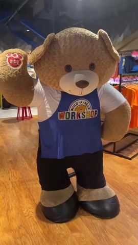 A mascot costume of a brown teddy bear stands in what appears to be a retail store. The bear costume is wearing a white t-shirt, a blue apron with a "Build-A-Bear Workshop" logo, and black pants. Its hands are large and brown, with one raised in a wave. The floor is made of wood. In the background, shelves are filled with colorful merchandise, including stuffed animals and clothing. Two individuals are visible in the background, out of focus. The bear costume is the primary focus of the shot.