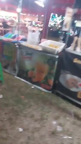 The video shows an outdoor market or fair at night. In the foreground, there are two food stalls. The stall on the left has a banner advertising "Jelly Ball" and appears to have colorful items on display. A green plastic stool is placed in front of this stall. The stall on the right has a banner with an orange design and text that appears to be "Es Jeruk Peras" (which translates to squeezed orange juice). There are oranges depicted on the banner. In the background, more stalls are visible with people working and browsing. Lights are strung up, suggesting a festive atmosphere. The ground is covered in grass and is littered with some debris, including paper and plastic wrappers. People are seen moving around and interacting at the stalls. The overall lighting suggests it is evening or night.