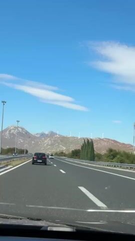 The video shows a view from a car driving on a highway. The highway has multiple lanes and is surrounded by a guardrail. The sky is blue with some white clouds. In the distance, there are mountains with a few wind turbines on top. There are other cars on the highway, including a black car in front of the camera vehicle. The time stamp indicates the video is from January 1st, 2023, starting at 00:00:00 and ending at 00:00:04.