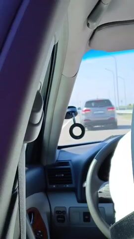 The video shows the interior of a car driving on a road. The perspective is from the passenger seat, looking forward. A gray SUV is visible in front of the car, also driving on the road. The sky is blue and the sun is shining. The interior of the car includes the dashboard, steering wheel, and part of the passenger seat. There is a circular object mounted on the windshield. The car appears to be moving at a moderate speed.