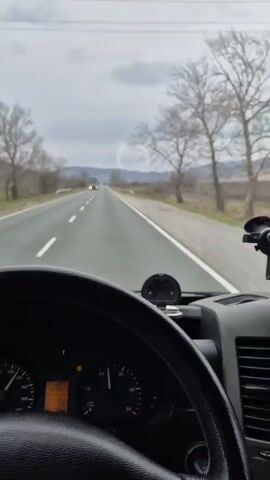 The video shows a dashboard-level perspective of a person driving on an asphalt road in what appears to be a rural area. The sky is overcast. The road ahead is mostly straight, with a distant truck visible in the oncoming lane. Two bare trees stand on the right side of the road, and fields and distant mountains are visible in the background. The dashboard contains a steering wheel, a car mount for a cell phone, vents, and a car radio. A "dp" branded air freshener hangs from the rearview mirror. A box of tissues rests on the dashboard. The camera view is focused on the driver's perspective of the road and surrounding scenery.