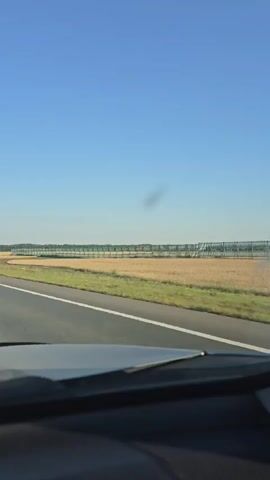 The video captures a first-person perspective from inside a vehicle driving on a paved road. The road has a white line marking the lane. To the right of the road is a vast, golden field, likely a crop, extending towards the horizon. In the distance, a line of green structures, possibly a fence or an irrigation system, runs parallel to the road. The sky is a clear, bright blue. At the beginning of the video, another car, a silver station wagon with a license plate beginning with "A910CV," is briefly visible in the right lane, overtaking the vehicle from which the video is being recorded. The front interior of the recording vehicle is partially visible in the foreground, showing the dashboard and windshield. There are no people visible outside the vehicles. The overall environment suggests a rural or agricultural setting under a clear, sunny sky.