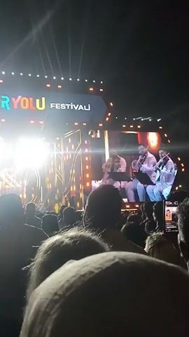 A video shows a live performance at the Güryolu Festivalı. The view is from the crowd, showing silhouettes of people with some hair visible. The stage is brightly lit and features a large screen displaying images of the performers. Three men are seated on the stage, singing into microphones. Behind them, the screen shows a full band performing. The background features vertical golden lines and decorative elements. Many in the audience are holding up their phones, presumably to record the event.