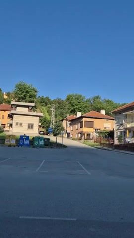 The video shows a quiet street scene in a residential area on a clear, sunny day. The camera pans slowly to the right, revealing several houses with red-tiled roofs, typical of a European village or town. In the foreground, a paved road with painted parking lines is visible. To the left, there are blue and green recycling bins. Behind the bins, a small, light-colored building stands. Further down the street, houses with yellow and white facades are seen, some with balconies and flower arrangements. Green trees and foliage line the background, suggesting a hilly or elevated terrain. The sky is a vibrant, cloudless blue. There are no people or vehicles visible in the frame, contributing to the sense of stillness and tranquility. The lighting indicates it is likely morning or late afternoon.