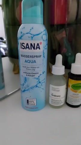 The video shows a group of beauty products on a white surface, arranged in a line. The products include a bottle of ISANA Aqua Water Spray, a bottle of The Ordinary Hyaluronic Acid 2% + B5 serum, a bottle of Alverde Vitamin C serum, and a box of SKIN1004 Madagascar Centella Hyalu-Cica Water-Fit Sun Serum SPF50+ PA++++. The background includes more beauty products and some other containers. The camera slowly moves from left to right, showing each item. There are no people in the video.