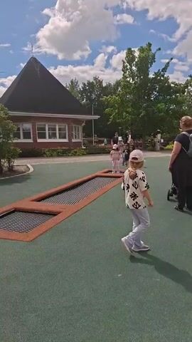 A group of children are playing on an outdoor trampoline that is built into the ground. The trampoline is rectangular with a brown frame. One child in a pink outfit and hat is jumping in the middle of the trampoline. Another child in a white shirt with a pattern and a pink hat is walking away from the trampoline towards the right side of the frame. Several other children and adults are visible in the background. There is a brick building with a dark, pointed roof behind the trampoline. The ground is covered in green turf. The sky is blue with white clouds. The video is shot from a low angle and is slightly shaky.