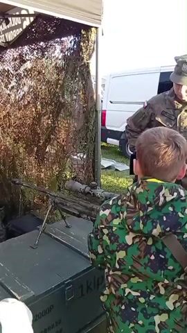 A person in a military uniform, wearing a camouflage hat, is demonstrating a sniper rifle with a scope and bipod. The rifle is placed on a dark green military-style box. A person in a camouflage jacket is standing to the right, watching the demonstration. The background shows a white van and an ATV. The environment appears to be outdoors, possibly at an event or exhibition.