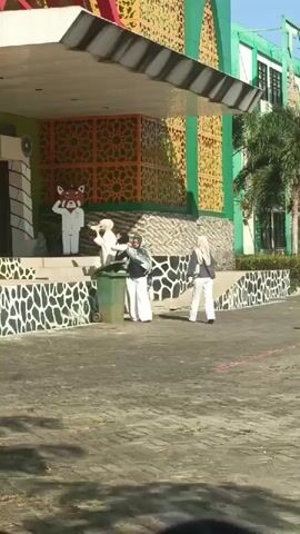A group of people are gathered outside a building. A person in a red panda costume is standing on a stage. Three other people in white outfits are standing near the red panda costume. Two people in white headscarves and white pants are walking away from the stage. A green trash can is located near the stage. The building has green and orange decorations on the facade. The ground is paved with asphalt and bricks. Sunlight is visible, indicating it is daytime.