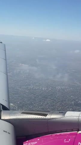 The video shows an aerial view from the window of an airplane. The wing of the plane is visible on the left side of the frame, and the engine is partially visible in the lower section. Below, a dense urban landscape stretches out, with buildings appearing as a uniform grey mass. A highway or road snakes through the city in the distance. The sky is blue with a few scattered clouds, and a haze obscures the far horizon. The overall impression is of a sprawling city viewed from a high altitude.