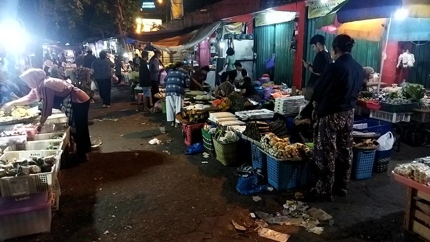 The video shows a busy night market scene. People are browsing and shopping among stalls filled with various goods, primarily food items. The stalls are illuminated by bright lights, creating a vibrant atmosphere. Some vendors are seen attending to customers, while others are arranging their products. The ground appears somewhat cluttered with discarded items, suggesting a lively and active marketplace.