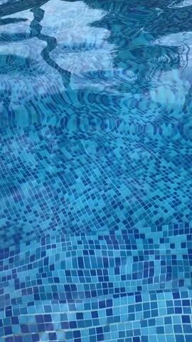 The video shows a close-up view of a swimming pool with a blue mosaic tile pattern at the bottom. The water is clear and ripples slightly, distorting the appearance of the tiles underneath. The camera remains static throughout the video, focusing on the play of light and water on the mosaic tiles. The pool steps are also visible.