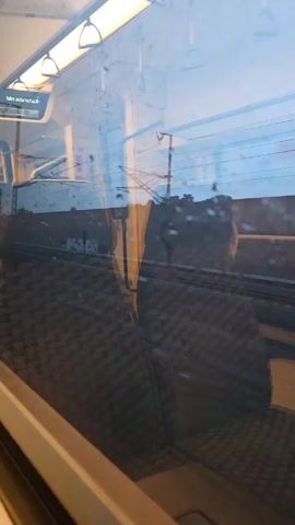 The view from inside a train carriage shows empty grey patterned seats next to a window. The window is slightly obscured by raindrops. Outside the window, railway tracks and overhead power lines are visible against a cloudy sky. The train appears to be moving. The carriage interior is lit with overhead lights.