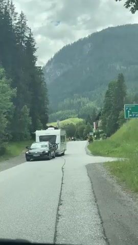 This video captures a scenic drive through a mountainous region, likely in Flachauwinkl, Austria. The scene is dominated by a paved road winding through a lush, green valley. On either side of the road, dense coniferous forests rise up towards imposing, mist-shrouded mountains. The weather appears overcast with dramatic cloud formations, suggesting a temperate climate.

The primary activity is a slow-moving vehicle procession. A dark-colored car is visible in the foreground, towing a white caravan. The caravan has a bicycle mounted on its rear. Further ahead on the road, a few figures, presumably people, are walking or standing near houses that dot the landscape. These buildings appear to be typical Alpine chalets, nestled into the hillsides.

Notable details include a green and white directional sign with the letter 'P' on the right side of the road, indicating a parking area. Another sign with Austrian flags is visible near one of the houses. The road itself shows signs of wear, with visible cracks and patches. The time of day is indeterminate due to the overcast conditions, but the overall atmosphere is one of a peaceful, rural journey. There is no strong emotional tone conveyed, rather a sense of calm observation of the passing scenery.