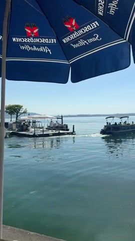 This video shows a view of a lake on a clear, sunny day. In the foreground, a blue umbrella with white lettering is visible at the top left of the frame, and a wooden surface is at the bottom right. In the middle ground, a dock extends into the water on the left, with a few people standing on it. A pontoon boat with several people on board is moving away from the dock, creating a wake. In the background, there is a distant shoreline with trees and buildings. The water is calm with some ripples. The sky is clear and blue.