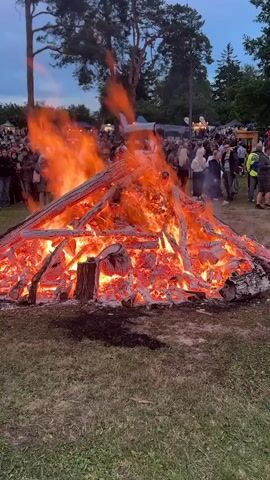 The video shows a large bonfire in a grassy field, with a crowd of people gathered in the background. The bonfire is made of large logs and branches, and it is burning intensely with bright orange flames and glowing embers. The fire is in the foreground of the video, taking up most of the frame. In the background, a crowd of people are standing and watching the fire. Some of them are dressed in casual clothing, and some are holding drinks. There are trees in the background, and the sky is a dark blue, indicating that it is dusk or evening. The overall atmosphere of the video is one of celebration and community.
