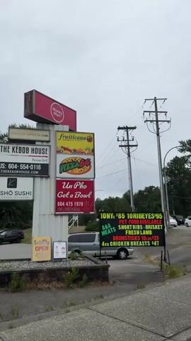The video pans across a parking lot in front of a strip mall. A white Jeep pickup truck is parked on the left side of the frame. A white Jeep Gladiator pickup truck is parked in the middle of the frame. Various signs are visible on a pole in the parking lot, including "Money Mart," "THE KEBAB HOUSE," "KOSHO SUSHI," "FRUITICANA," and "Un Pho." Other businesses in the strip mall include "MEATS" and "PITA PAN DONAIR." Several cars are parked in the lot. Trees and power lines are visible in the background. The sky is overcast.
