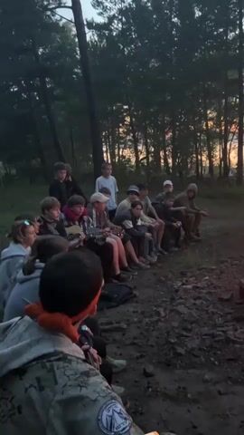 A group of children and young adults are gathered around a campfire in a wooded area. The sun is setting, casting a warm glow over the scene. The individuals are sitting on the ground or on logs, facing the fire. Some are looking at the fire, while others are talking to each other. The atmosphere appears relaxed and communal.