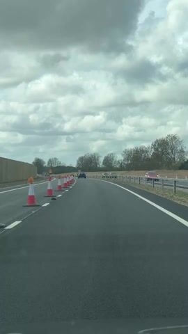 The video shows a view from a car driving on a highway. The day is overcast, with large, fluffy clouds filling the sky. The road is lined with red and white traffic cones, indicating a lane closure or construction zone. Several cars are visible ahead, driving in the open lane. There is a guardrail on the right side of the road and a concrete barrier on the left. Beyond the guardrail and barrier, trees and foliage are visible. The asphalt of the road looks new, with fresh white lane markings. A sign with an arrow indicating a lane change is visible between the traffic cones. The camera seems to be mounted inside a car, offering the perspective of the driver. Music plays in the background.