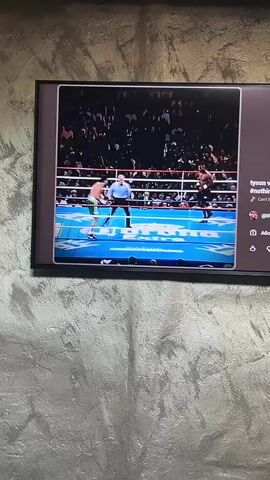 A television screen, framed in black, is mounted against a textured, light gray wall, displaying a boxing match. The video on the screen shows a boxing ring with red ropes and a blue canvas, on which the text "IRON TYXON" is visible. Two boxers are engaged in the fight; one wears black shorts and black gloves, while the other is in green shorts and red gloves. A referee in a white shirt and black pants is also present in the ring. A large crowd of spectators can be seen in the background of the arena.

The video appears to be from a streaming platform, possibly YouTube, as indicated by the interface elements visible to the right of the video frame. These elements include the title "tyson vs mcneely," hashtags like "#nothingimposs" and text "Can't be touched," and a partially visible username "@IRONTYXON3." Below these, icons for sharing and other functions are visible, along with the word "Абониране," which is Bulgarian for "Subscribe." The video itself shows different moments from the boxing match, suggesting a highlight reel or a montage of the fight's action. The overall scene captures the intensity of a professional boxing event being viewed in a domestic setting in Kozloduy, Bulgaria.