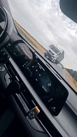 The video shows a close-up view of a car's dashboard, specifically the digital infotainment screen. The screen displays song information for "Beauty and the Beast" and playback controls. A steering wheel is partially visible on the left. Outside the car, a wide, flat landscape, possibly a road or airfield, is visible under a cloudy sky. The camera is tilted, giving the impression of movement.