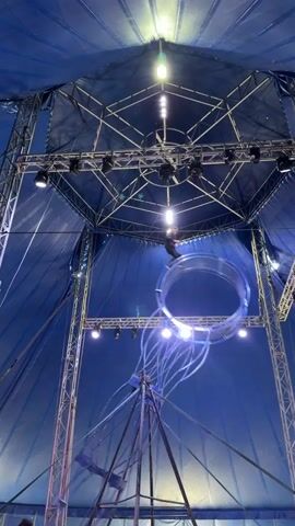 A person is suspended high above an audience inside a blue circus tent. They are performing a stunt with a large circular apparatus that appears to be made of metal and some kind of translucent material. The apparatus is suspended from the tent ceiling by cables and beams, and the performer is either on top of it or inside it, moving as it rotates or swings. Lights are positioned around the apparatus, illuminating the performance. The audience is seated below, watching the act.