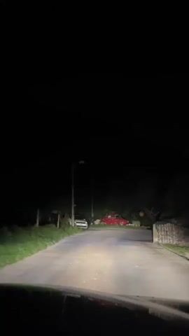 The video shows a dark night scene with headlights illuminating a rural road. Two cars are visible: a white car parked on the left side of the road and a red car parked further down on the right. The red car appears to be an older model, possibly a Volkswagen Beetle, and is decorated with lights or reflectors. Trees and vegetation line the sides of the road, and a street lamp is visible. The perspective is from inside a vehicle, suggesting the video is being recorded from a moving car. The atmosphere is quiet and still.