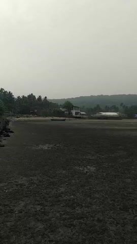 The video shows a wide, empty beach on a cloudy day. The beach is mostly flat and covered in dark, muddy sand, with some areas of standing water visible. In the distance, there is a line of trees and some buildings along the shoreline. The sky is overcast and gray.  A small boat is visible, off to the left side of the frame, in the mid ground. The camera slowly pans from left to right.  There are no people visible in the video.