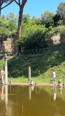 The video shows a serene scene at Villa Adriana, with a calm body of water reflecting the ancient architecture and statues. Several people are walking along a pathway adjacent to the water. The architecture includes Roman columns, arches, and remnants of a large building on a small hill in the background. The water is an amber color, and the reflections are distinct. The landscaping includes trees and grass. The sky is blue with light clouds.