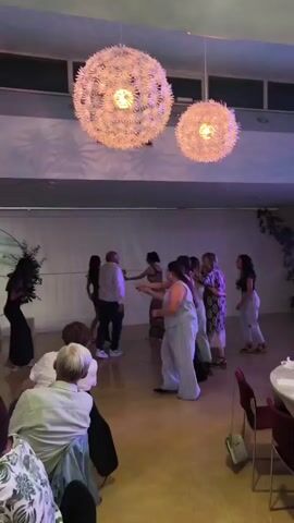 A group of people are dancing in a room. Two large, spherical light fixtures hang from the ceiling, emitting light. The walls are a light blue, and a green leafy plant is visible on the right side of the room. Some of the dancers are wearing formal attire, such as dresses. There are tables with chairs set up in the foreground.