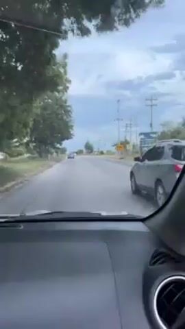 The video shows a view from inside a car driving on a road. There are two other cars ahead. A rainbow is visible in the sky. The landscape includes trees, telephone poles, and some buildings in the distance.  The sky is cloudy. The road appears to be paved and has some cracks.