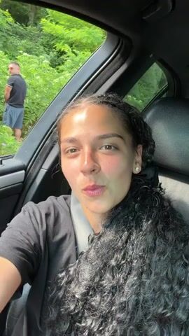 The video begins with a close-up shot of a young woman with curly dark hair, wearing a black t-shirt, looking at the camera and smiling. In the background, through the car window, a man in a black t-shirt and blue shorts is visible urinating by the side of a road, surrounded by lush green foliage and some purple flowers. The woman turns to face the side, and the camera follows her movement, showing her arm with several bracelets. The scene then shifts to a wider view of the man in the background, who is now a bit further away and angled away from the camera. The video continues with a glimpse of the car interior and the surrounding greenery. The overall impression is a casual, candid moment captured during a car ride in a natural, outdoor setting. The time of day appears to be daytime, with bright lighting suggesting sunny weather. The location context provided is Garnia, Bulgaria. There is no visible text in the video.