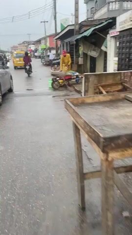 The video shows a man cleaning produce on a rainy street. The man wears a yellow shirt and a red baseball cap. He is using a rag and a container to wash vegetables on a small cart with three wheels. The cart also holds other items, including what appear to be bananas. The street is wet, and there are buildings and other people visible in the background, including a motorcycle and a yellow cab. There are various stalls or shops nearby.