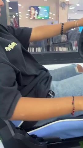 A woman with tan skin and dark hair is playing a racing video game. She is sitting in a racing seat with a steering wheel, gear shift, and pedals. The screen shows an orange Corvette racing on a track. She is wearing a black t-shirt, ripped jeans, and several bracelets on her wrists. The game is displayed on a large flat screen TV. The environment looks like a store with display cases and lights overhead.