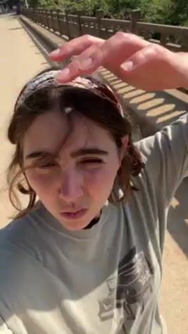 A woman films herself in an outdoor setting. She is standing on a bridge with a carved railing, and she appears to be using a front-facing camera, as her arm is visible in the frame. She wears a gray shirt and a headband. Behind her, there are mountains covered in trees. Below, water can be seen. In the video, she adjusts her hair and sticks her tongue out.
