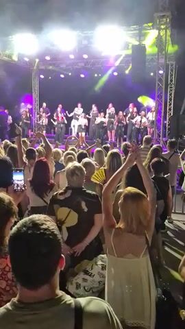 The video shows a live band performing on a stage at night. The stage is lit with purple and green lights. The band consists of many members, some playing instruments and others singing. In front of the stage, a crowd of people is dancing and enjoying the performance. Some people in the crowd are holding up their phones to record the event. The overall atmosphere appears to be festive and energetic.