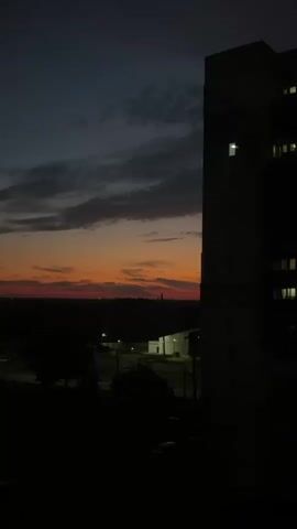 This is a vertical, silent video clip featuring a dusky sky with clouds painted in hues of orange, red, and deep blue. The sun has likely set, casting a warm glow on the horizon. In the foreground, a large, dark silhouette of a building occupies the right side of the frame, with a few lit windows visible. Below the horizon, the landscape is shrouded in darkness, with the faint outlines of trees and some distant lights suggesting human habitation or structures. The overall impression is one of twilight, with the last remnants of daylight fading into night. There are no people or discernible actions in the video.