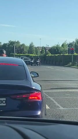 The video shows a blue car stopped at an intersection, waiting for the light to change. Two motorcyclists are stopped across the intersection, also waiting for the light. The light turns green, and the motorcyclists drive forward. The scene is recorded from inside the blue car. The weather appears sunny and clear.