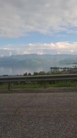 The video shows a view from a moving vehicle, looking out at a large body of water, possibly a lake, bordered by rolling hills and mountains. The sky is partly cloudy with patches of blue. In the foreground, there is a road and a metal guardrail. Vegetation, including green grass and some bushes, is visible between the road and the guardrail. The mountains in the distance appear to be hazy or covered in atmospheric perspective. There are some structures visible on the far shore of the water, suggesting a settlement or industrial area. A tall pole, possibly a street light, is seen in the middle ground on the right side of the frame. The overall impression is a scenic landscape viewed during daytime.