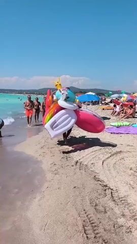 A man walks along a sandy beach carrying a stack of colorful inflatable pool toys. The beach is crowded with people sunbathing and swimming in the clear blue ocean under a bright sunny sky. Some people are walking along the shore, while others are in the water. In the foreground, a woman wearing a bikini is walking away from the camera, and another person is partially visible behind her. The waves are gently lapping at the shore.