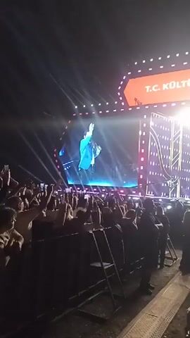 From a low angle, a video captures a crowded outdoor concert at night. Attendees, mostly silhouetted, are facing a large stage. On the stage, the band is visible in the background, with the image of a man in a blue jacket projected on the big screen behind them. The stage design features pink and white light elements, and the logo of the event, 'SK', is prominently displayed. The banners above the stage read 'T.C. KÜLTÜR VE TURIZM' and 'TÜRKIYE KÜLTÜR'. Many audience members hold up their phones, presumably recording the performance. A low barrier stands in the foreground, separating the cameraman from the crowd.
