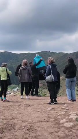 The video shows a group of approximately six adults gathered on a dirt and rocky elevated area, likely a hilltop or ridge, in Jeti-Ögüz, Kyrgyzstan. In the center, one individual wearing a bright blue jacket is being assisted by another person to mount a dark horse. Several other adults stand around, observing the activity. A dog is visible on the far left of the frame. The background features rolling green mountains and hills under an overcast, grey sky. A small village or settlement is discernible in the valley below. The scene occurs during daytime with cloudy weather. No visible text is present in the video.