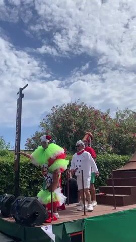 A small group of people are on a decorated float, possibly for a parade or festival. A person in a green and red layered costume, resembling a tutu, and a Santa hat, is on the left side of the float. Next to them is a person in a chef's costume and another in a red shirt and green shorts. At the back of the float is a large, ornate structure that appears to be a volcano or a similar monument. A statue of a woman with elaborate headwear stands on top of this structure, and smoke or mist is emanating from it. The float is decorated with green and red fabric. In the background, there are trees with pink flowers and a bright blue sky with white clouds. Two large black speakers are visible on the left side of the float.