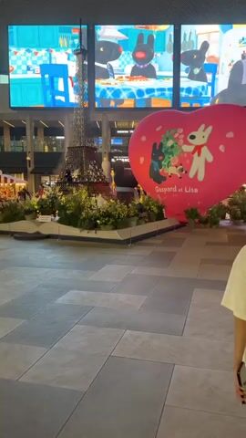 The video pans across a mall interior. In the foreground, large grey and beige square tiles form the floor. A woman with short dark hair, wearing a white t-shirt and white shorts, walks away from the camera. To her left is a large pink heart-shaped inflatable with cartoon characters on it, and behind that, a miniature Eiffel Tower replica surrounded by greenery. Above them, a large screen displays animated cartoon characters in a kitchen. To the woman's right, a red stall with signage is visible, with people interacting at the counter. Further in the background, other people are seen walking and interacting in the mall. The mall features multi-story architecture with arched windows and a high ceiling with visible structural elements.