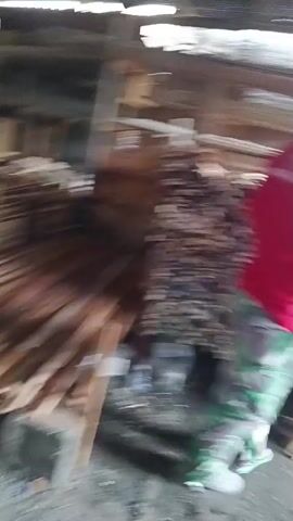 A man wearing a red shirt, camouflage pants, and a cap is carrying a piece of wood in what appears to be a woodworking shop or lumberyard. He is walking away from the camera, heading towards the right side of the frame. The environment is cluttered with stacks of lumber of various sizes and shapes. Some of the wood is piled high against a wooden structure, while other pieces are laid out on the floor, supported by bricks. The ground is dirt or gravel. The lighting suggests an indoor space, possibly with natural light coming from an opening or windows not visible in the frame.